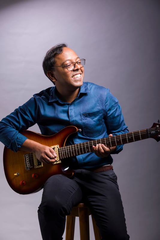 Bijoy Thangaraj seated and smiling while holding an electric guitar